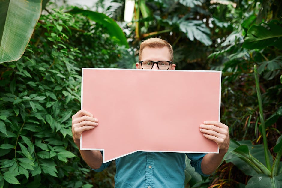 Man in glasses holding a pink speech bubble sign in lush greenery setting.