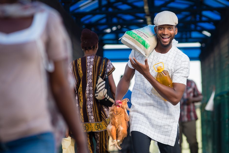 Man carrying groceries and a chicken in an outdoor market, smiling happily.