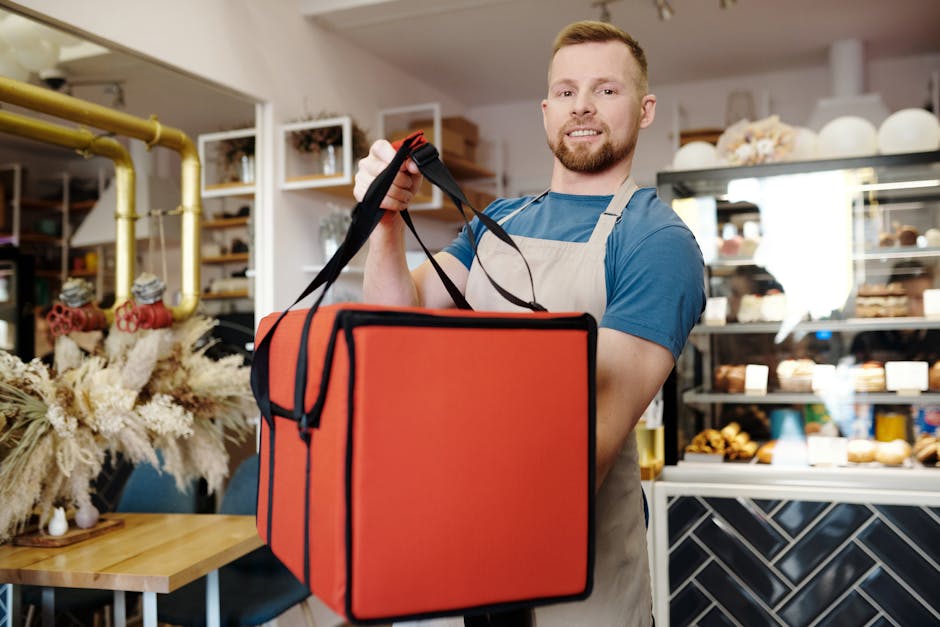 Smiling man in an apron holding a delivery bag in a cozy cafe setting, ready for service.
