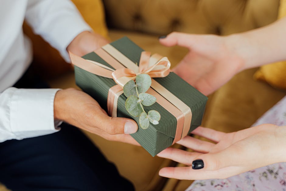Two hands exchanging an elegantly wrapped green gift box with ribbons and foliage.