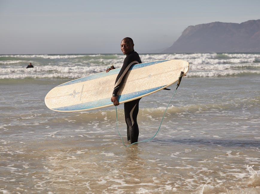 A confident surfer holding a surfboard in the waves with a scenic beach backdrop.