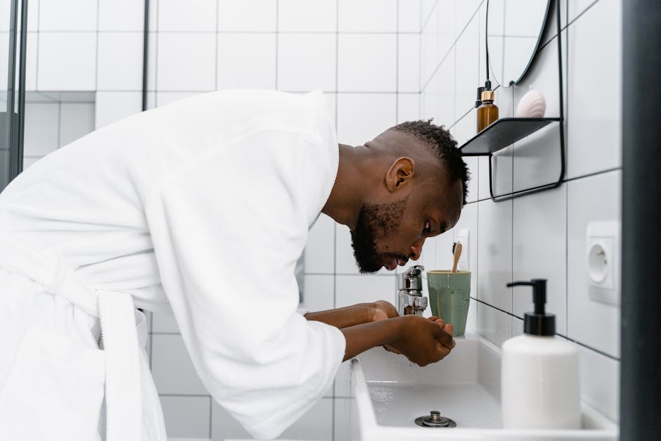 Young man in bathrobe washing face in a modern white bathroom.