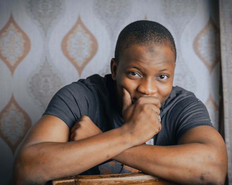 A smiling man leaning on a chair in a cozy room with patterned wallpaper.