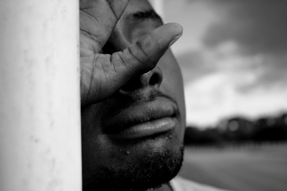 Grayscale close-up of a man outdoors, exhibiting a pensive expression, against a cloudy sky.