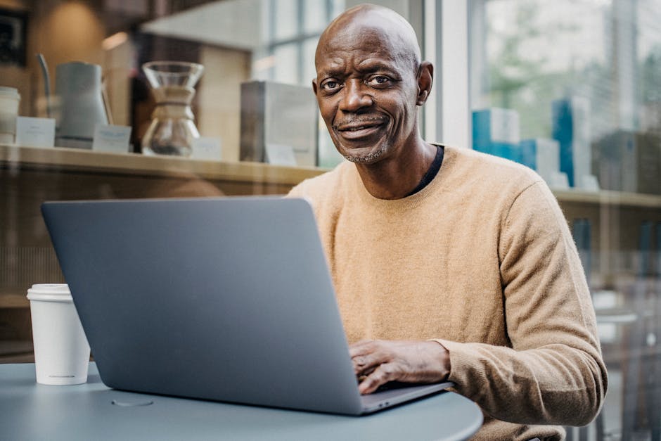 Positive mature bald African American male freelancer smiling and looking at camera while working on laptop sitting in cafe with cup of takeaway coffee